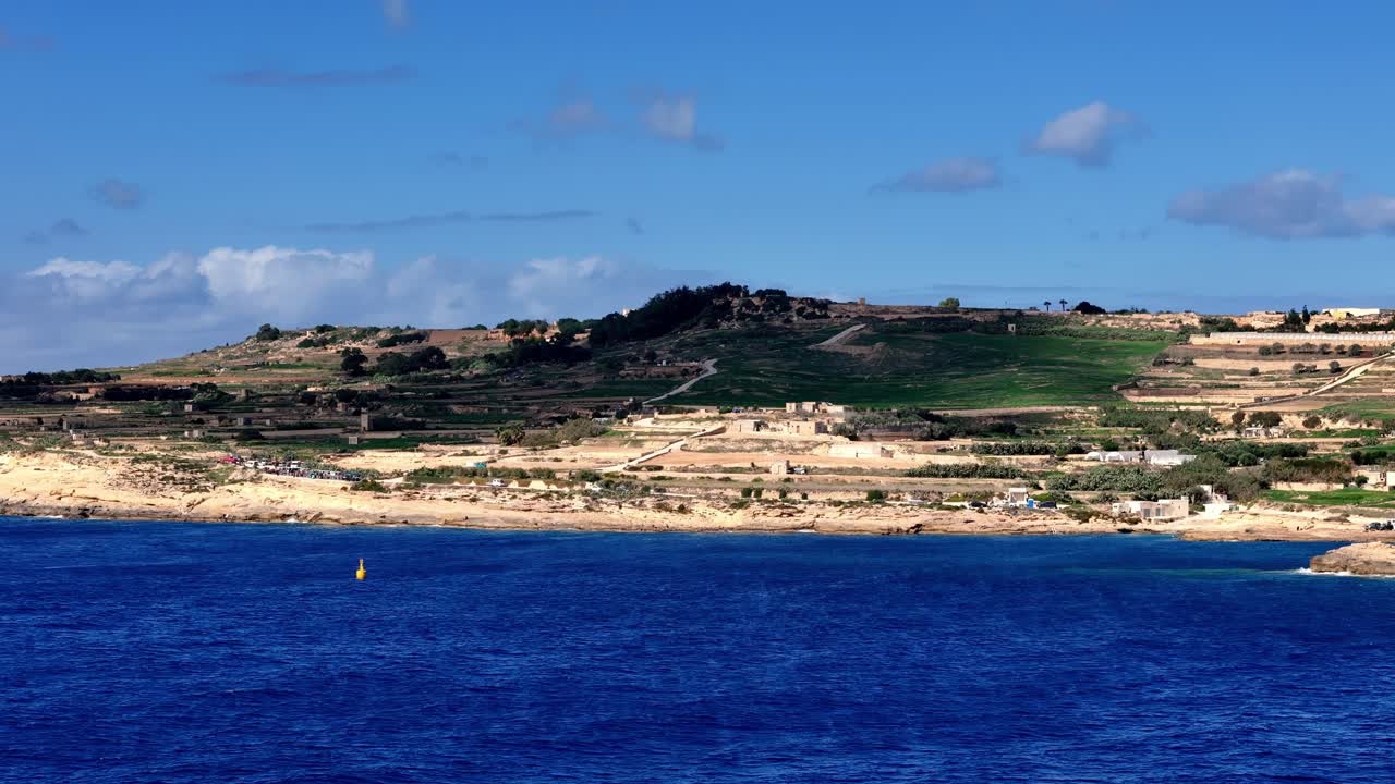 Vivace veduta grandangolare di scogliere calcaree terrazzate baciate dal sole a Malta durante una limpida giornata estiva, con acque mediterranee di un blu intenso sotto un cielo azzurro brillante con nuvole bianche sparse.
