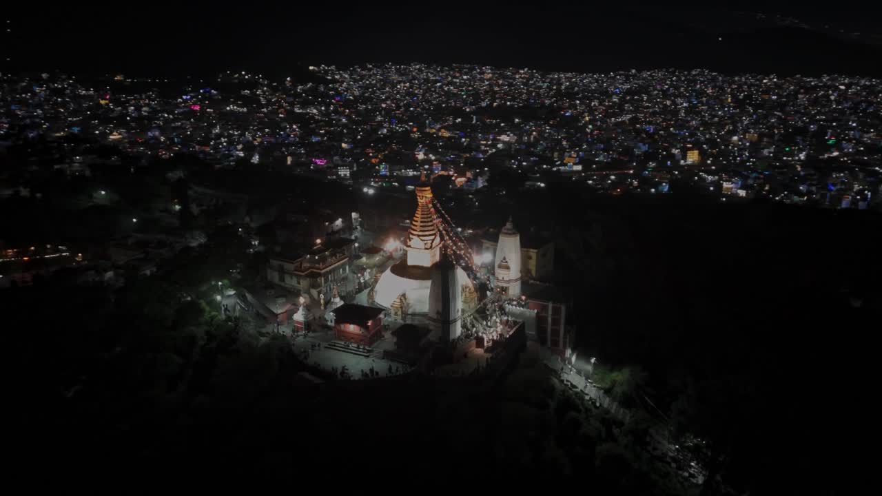 Aerial night drone footage of Swayambhunath Stupa in Kathmandu, glowing with warm lights atop the hill, showcasing its spiritual ambiance, iconic Buddhist architecture