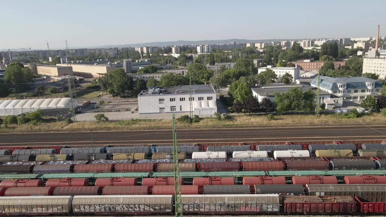 Aerial view of a busy rail yard filled with multiple freight train wagons, including tankers, hoppers, and open cars. The industrial setting includes nearby buildings and warehouses.