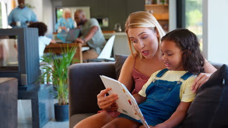 Madre-E-Hija-Leyendo-Un-Libro-En-Casa-Junto-Con-Una-Familia-Multigeneracional-En-Segundo-Plano