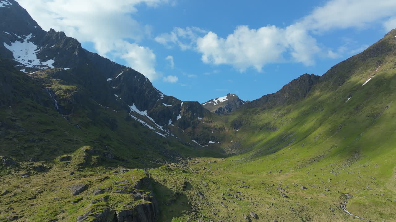 Verdant valley nestled between snow capped mountain peaks, revealing pristine Lofoten landscape under bright summer sunlight with sweeping Norwegian wilderness scenery