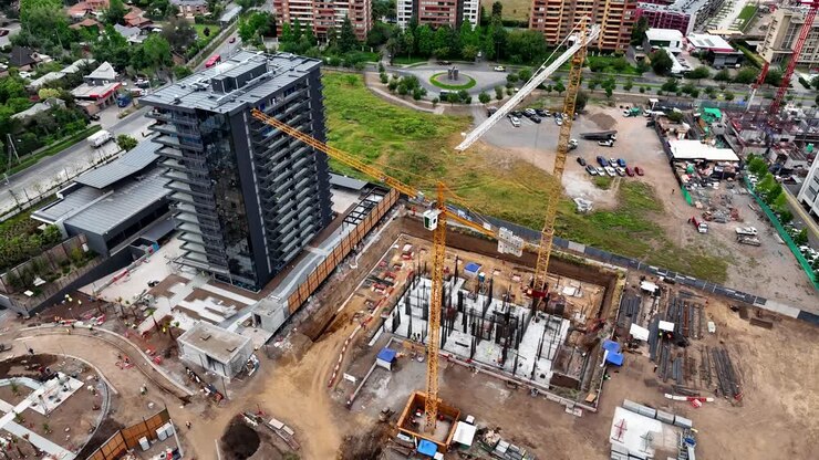 Vista aérea descendente con dron de una obra de construcción urbana con grúas amarillas, torres residenciales y progreso de desarrollo con colinas de fondo, Chile