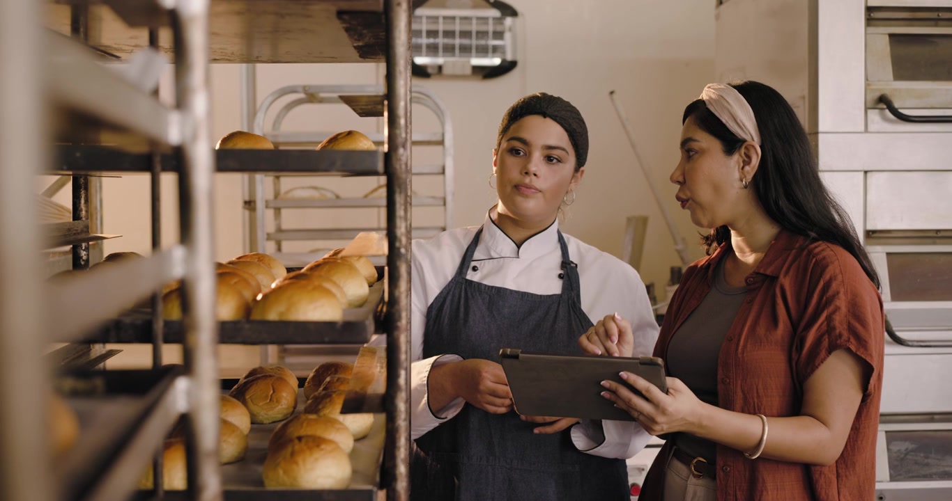 Chef e manager donne di diverse etnie che ispezionano pagnotte fresche sulle griglie in una panetteria, confrontando i dati su un tablet.