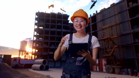 trabajadora asiática con gafas de protección y casco de seguridad sonriendo y señalando una tableta en su mano mientras está de pie en el sitio de construcción