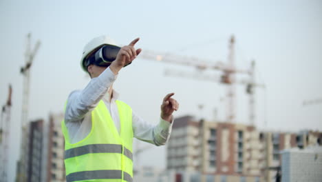 Portrait-of-a-male-inspector-analyzing-the-work-of-builders-using-virtual-reality-glasses.-A-man-in-a-helmet-and-a-protective-vest-stands-in-VR-glasses-and-moves-his-hands.