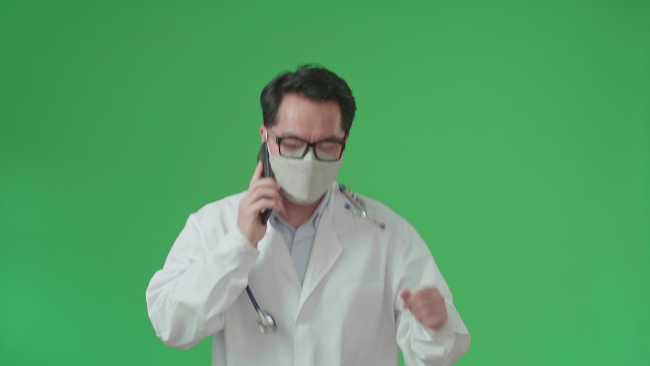 The Front View Of Asian Doctor With Stethoscope Wearing Mask, Glasses, And White Coat Talking On Mobile Phone While Running On Green Screen Chroma Key.