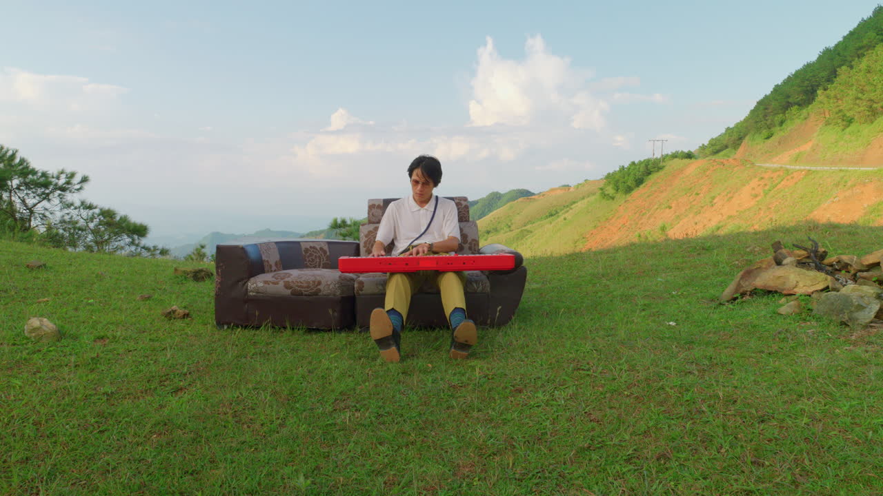 artist play piano keyboard while sitting on outdoor leather vintage couch placed outside in mountains valley