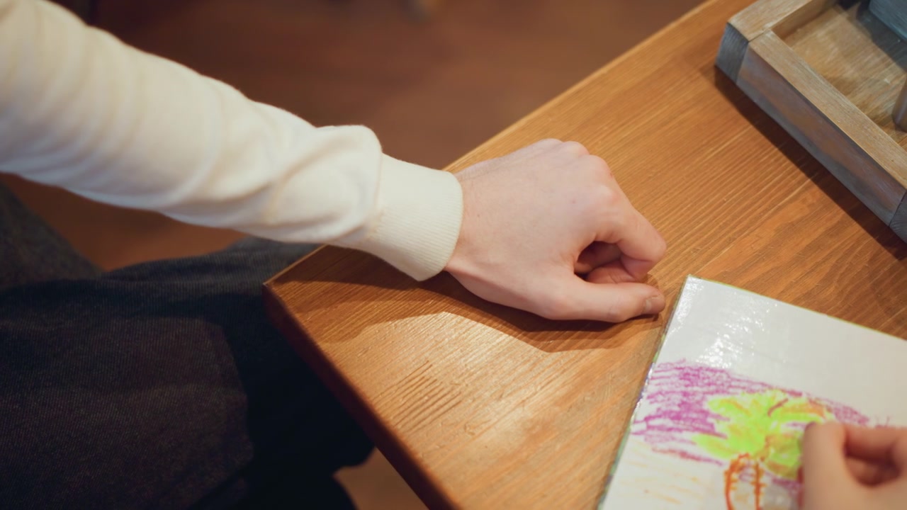 Close up of two people one adult and child interacting with colored pencils on paper at wooden table. visible hand movements show process of drawing or coloring together in indoor setting