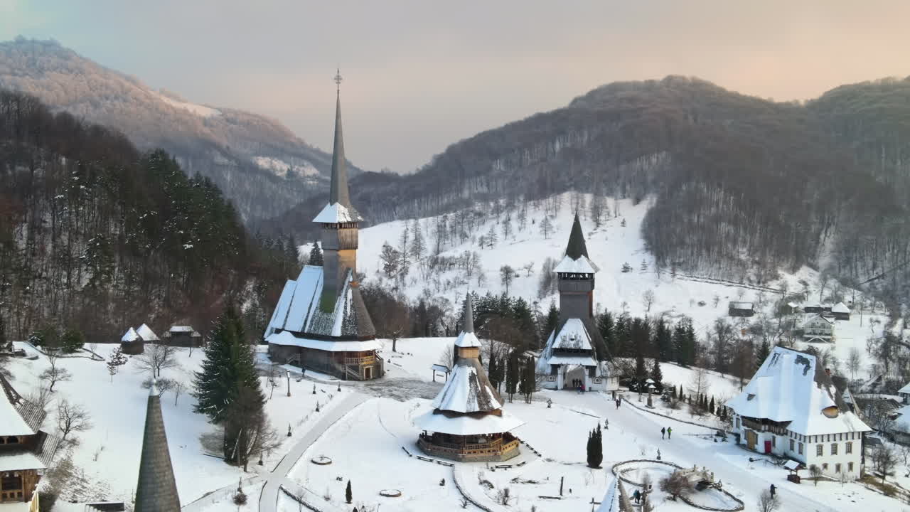 Lot z drona nad monastyrem Barsana w Rumunii. Główny kościół i inne budynki, goście, wzgórza pokryte nagim lasem i śniegiem, zima