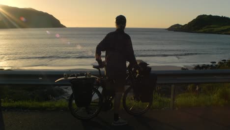 young male arriving at a beautifull veiwpoint over a beach and the sea with fully packed touring bike in sunset