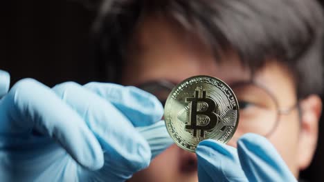 asian man in gloves carefully examines a gleaming golden bitcoin cryptocurrency