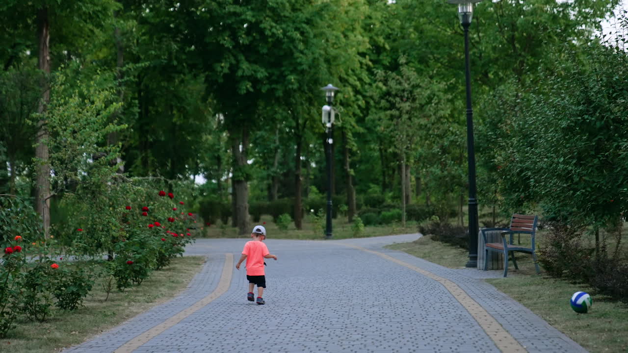 Rear view of a little toddler boy running away from camera. A ball rolls behind the kid by the park.