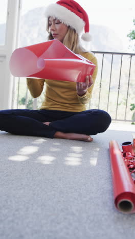 vertical video of biracial woman wearing santa hat packing christmas presents, slow motion