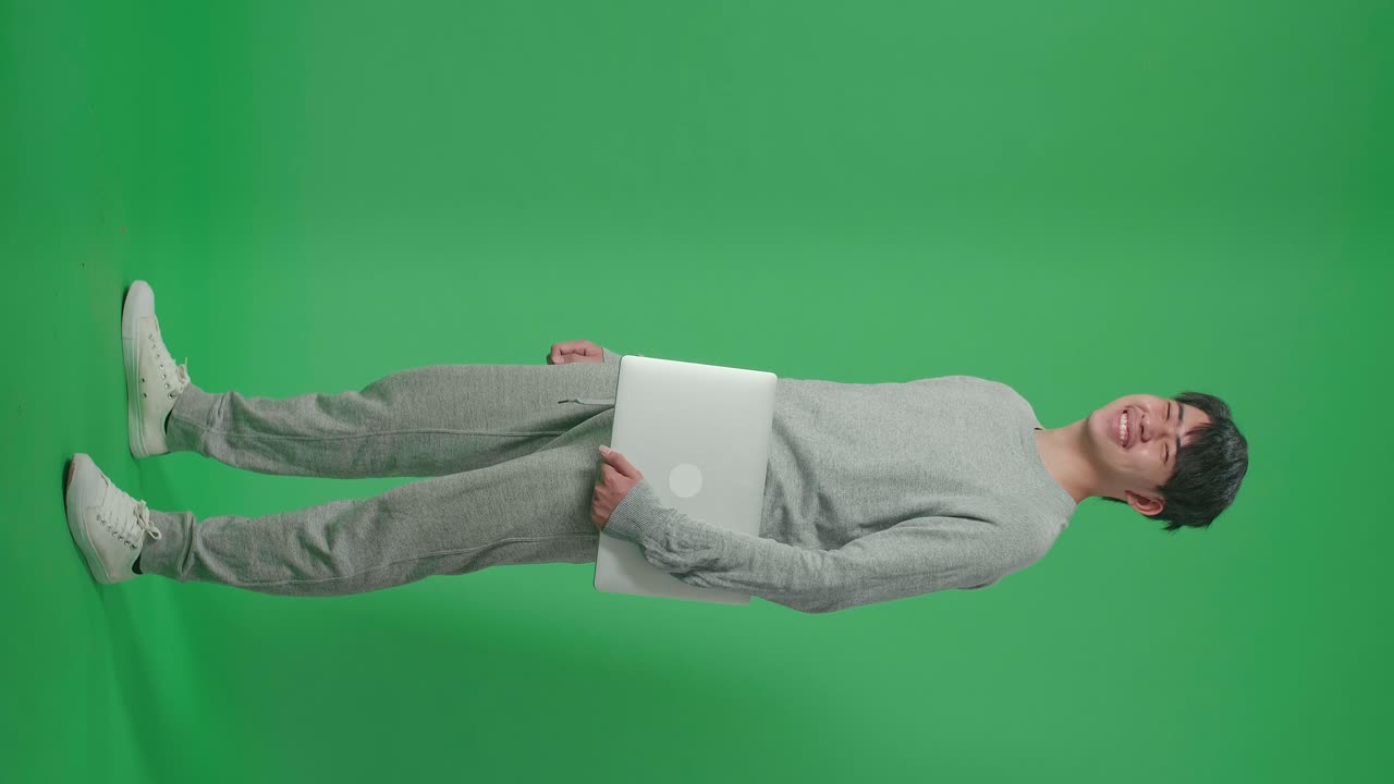 Full Body Of Side View Asian Man Holding Computer While Standing In Front Of Green Screen Background