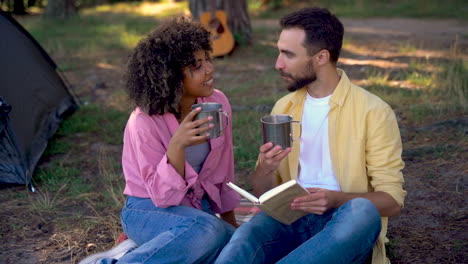 Happy-hiker-couple-camping-in-the-forest.-Caucasian-man-reads-book,-young-black-female-gives-him-a-steel-cup-of-coffee.