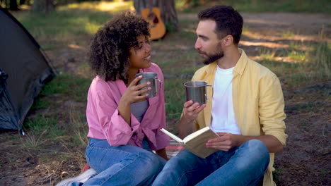 happy hiker couple camping in the forest. caucasian man reads book, young black female gives him a steel cup of coffee.