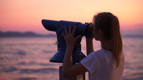 una hermosa joven mirando al cielo con un telescopio de metal en la costa durante la puesta de sol - primer plano