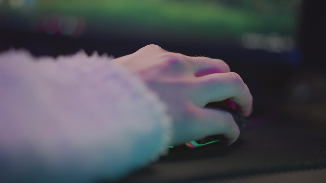 Close up hand of person in fluffy sweater resting on mouse in front of monitor, illuminated by subtle pink and green ambient lighting, relaxed mood, modern workspace environment