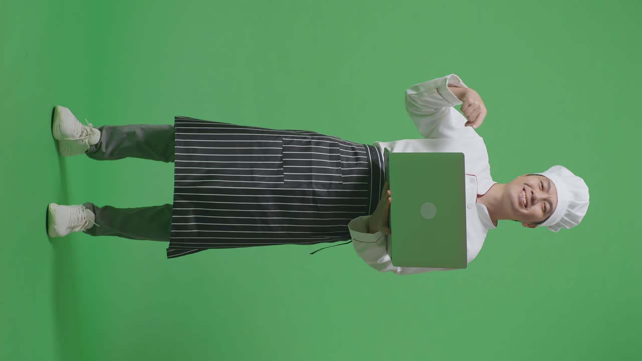 cuerpo lleno de hombre asiático chef sonriendo y señalando a una computadora portátil en la mano mientras estaba de pie en el estudio de fondo de la pantalla verde