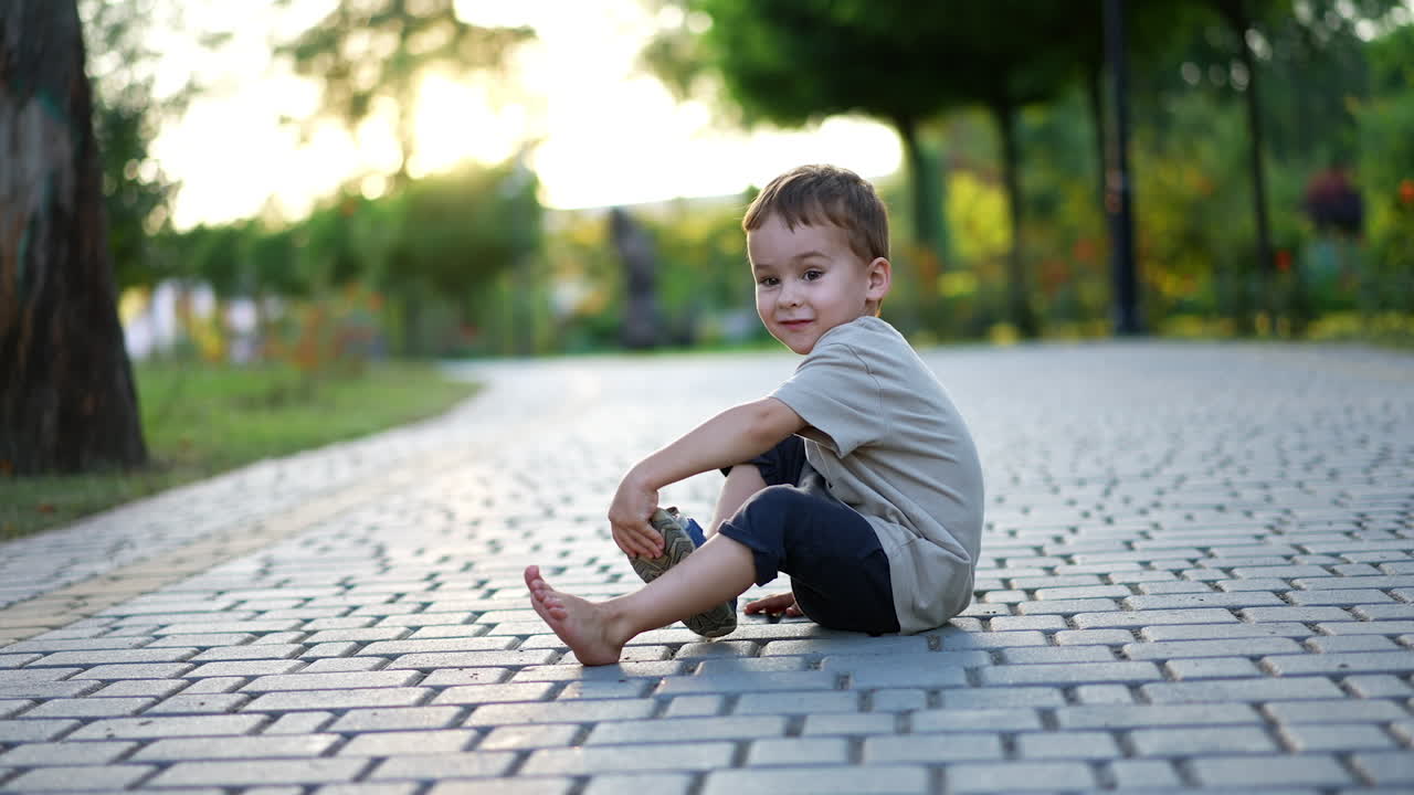Cute little toddler boy sits on the wide paved alley. Kid takes off his shoe and throws in the direction of camera. Blurred backdrop.