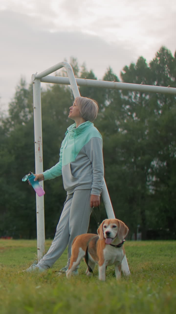 Una mujer blanca entrena a un beagle en un campo mientras sostiene una botella; escena de adiestrador canino y entrenador a tiempo parcial, sudadera con capucha color menta, fondo con postes de portería, práctica de obediencia lúdica, rutina de ejercicios por la tarde y vínculo afectivo con la mascota