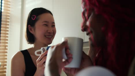 Two-beautiful-women-multiethnic-friends-drinking-coffee-while-talking-in-the-kitchen-at-home,-close-up