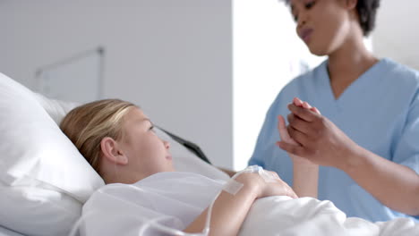 diverse female doctor and girl patient lying on bed holding hands in hospital, slow motion