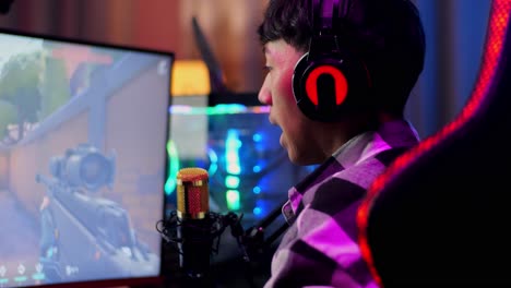 vista de atrás de un niño asiático que juega un juego a través de la red en una computadora personal, hablando con los participantes en el micrófono. videojuego de transmisión en vivo, escritorio iluminado por luz de tira de led rgb