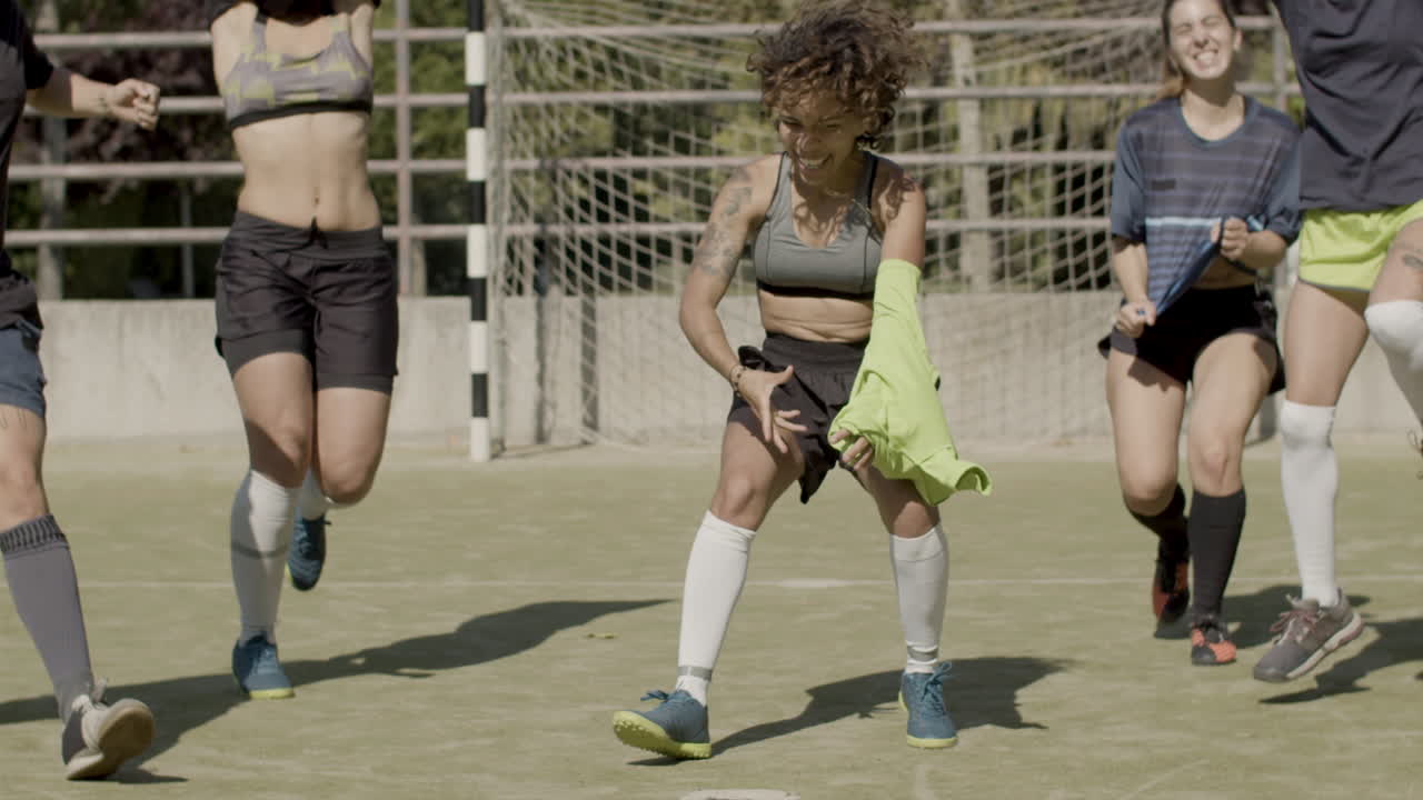 달리고, 티셔츠를 벗고, 카메라를 바라보며 승리를 기뻐하는 명랑한 여자 축구 선수들의 앞모습