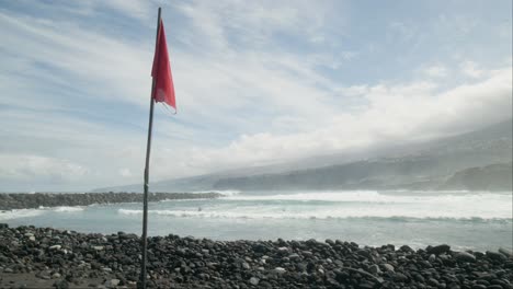 red warning flag with slowmotion group of surfers surfing on ocean waves on pebble beach, playa martiánez, puerto de la cruz, tenerife, canary islands in spring