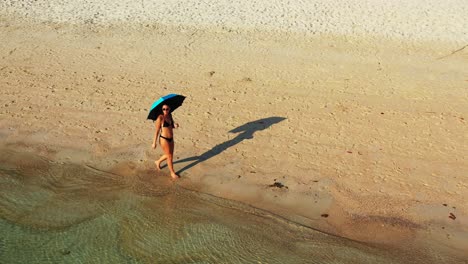 Girl-with-azure-umbrella-walking-barefoot-in-swim-suit-on-sandy-beach-washed-by-clear-emerald-water-of-turquoise-lagoon-in-Fiji