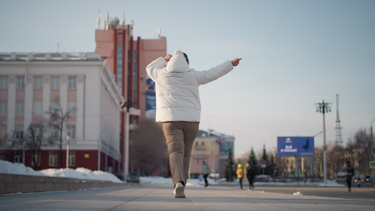 Back view choreographer walking down tiled path beside staircase on busy urban street during winter wearing puffy coat beanie surrounded by snow piles billboard cars pedestrians and city buildings