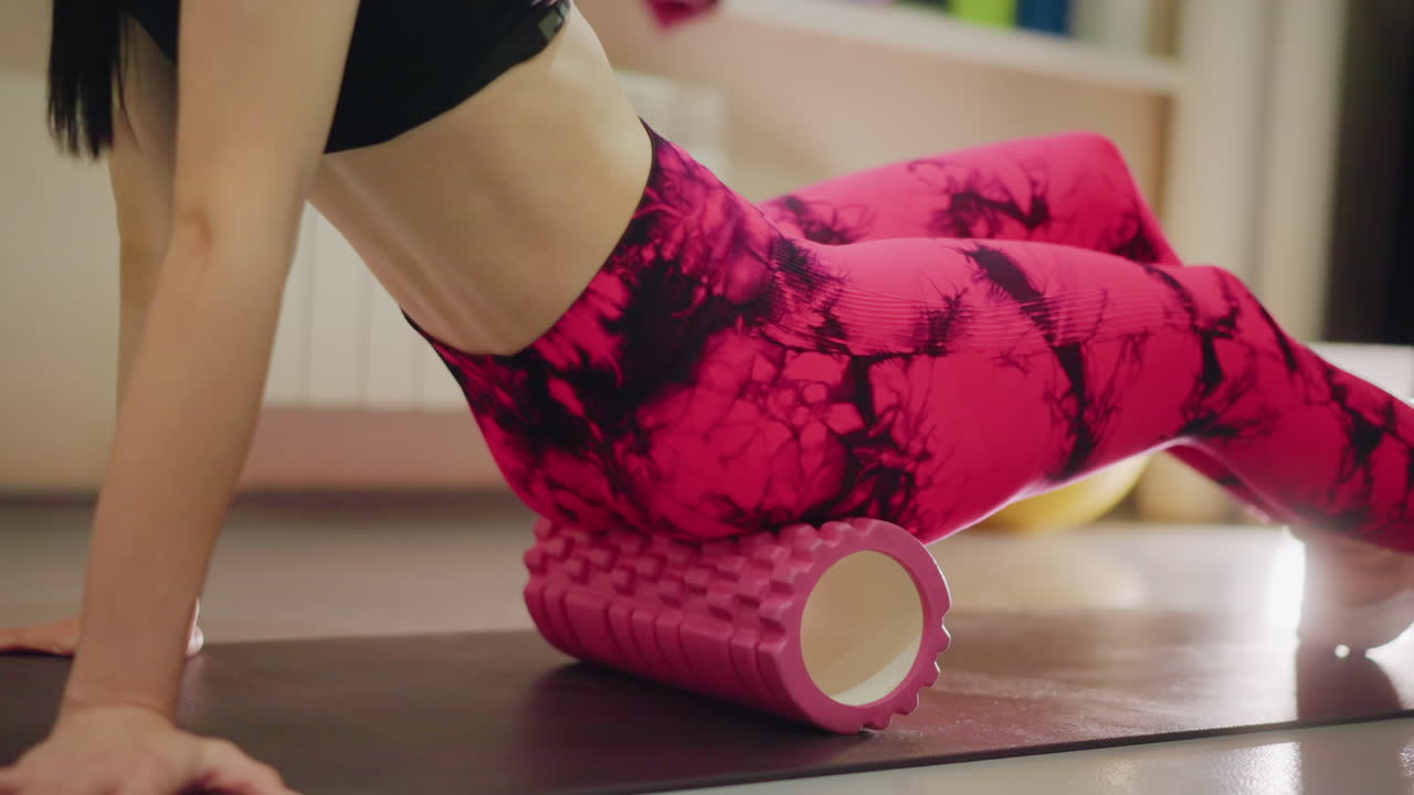 woman on mat using foam roller on glute, hands bracing on floor, pink patterned leggings and black sports bra, slow controlled roll for tension release, soft indoor light, calm recovery atmosphere