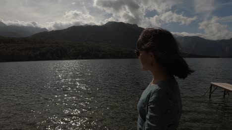 A-young-woman-with-brown-hair-and-sunglasses-walking-along-a-pond-along-the-lake