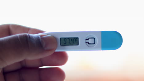 ill man holds digital thermometer with body temperature reading in fahrenheit up close to the camera against white background