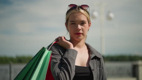 white woman in grey shirt and sunglasses over her head carries colorful shopping bags over her shoulder, with a focused expression, with an electric pole blurred in the background