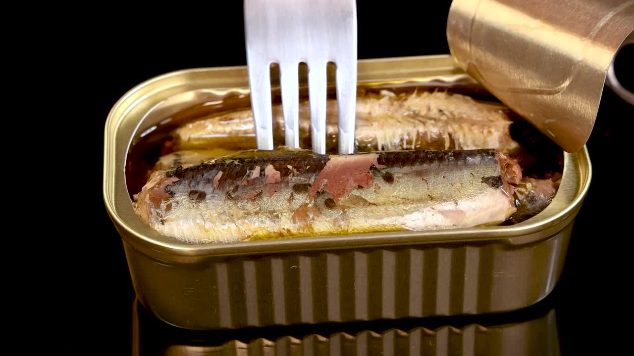 A metal fork lifts a sardine from an open tin can filled with oil, under bright studio lighting, against a black background with minimal reflections