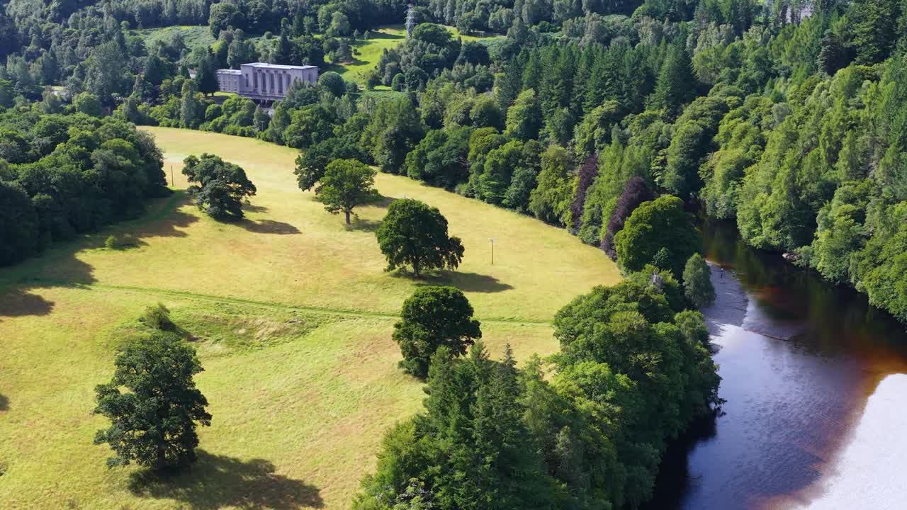 Drone glides above lush river bend, hydroelectric station, green forest, and sunlit landscape