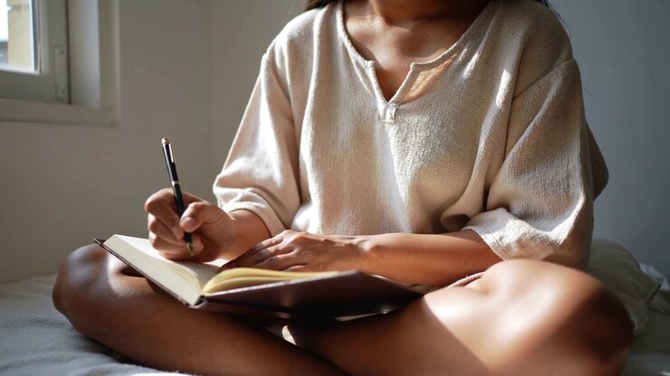 Sunlight falling from window starting woman journaling in longsleeve on bed, with hardback book