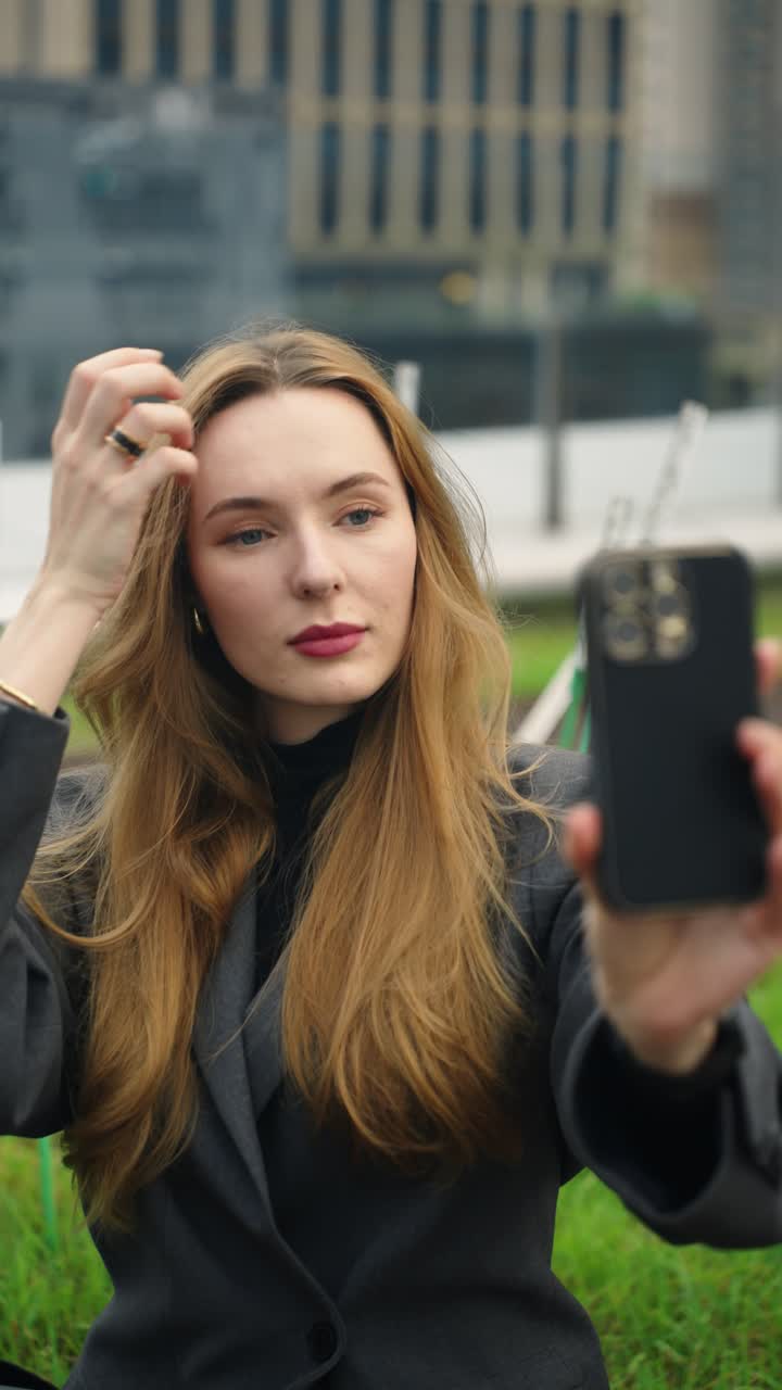 Fashionable businesswoman with long hair in a dark blazer styles her hair and takes a selfie with smartphone on a modern city terrace - parallax slow motion