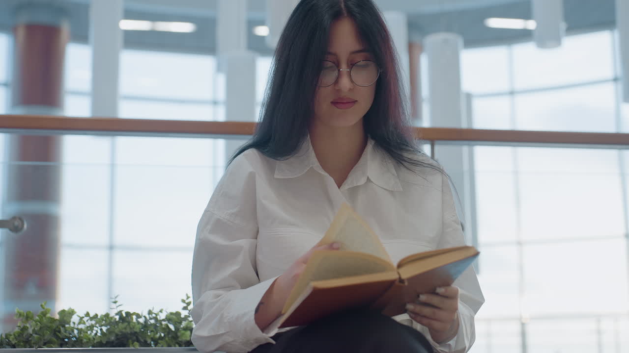 Young woman seated indoors gently flips through book with calm expression, surrounded by greenery and modern glass panels, absorbed in thought with soft lighting creating peaceful atmosphere