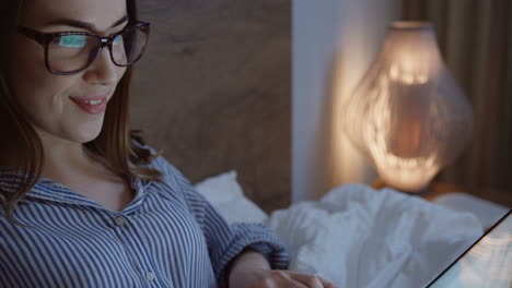 close-up view of the smiling caucasian young woman typing on the keyboard of the laptop late at night in the bed