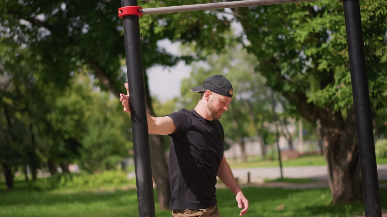 Caucasian Man Warming Up At Outdoor Bars. Park Coach Prepares For Calisthenics Session, Adjusting Grip On Horizontal Bar, Focused Expression, Leafy Background, Wearing Backward Cap And Cargo Pants