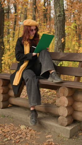 young artist deeply focused on sketching seated on wooden park bench in peaceful autumn park, one leg on bench, one on ground, with bag on floor, surrounded by golden leaves and serene nature