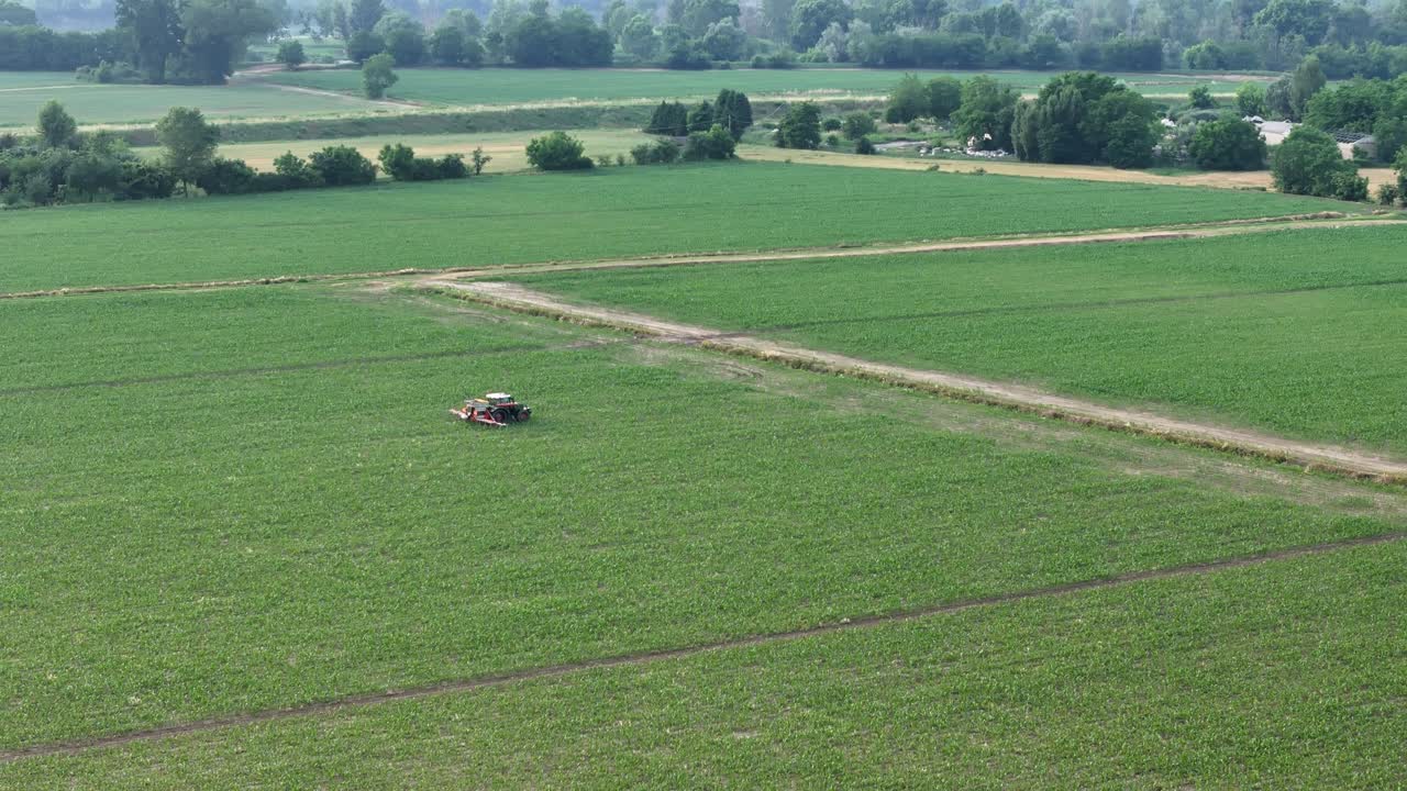 Real-time drone orbit around a tractor with boom sprayer operating in a green maize field close to rural houses, capturing the precise movement of crop treatment and the vibrant countryside landscape