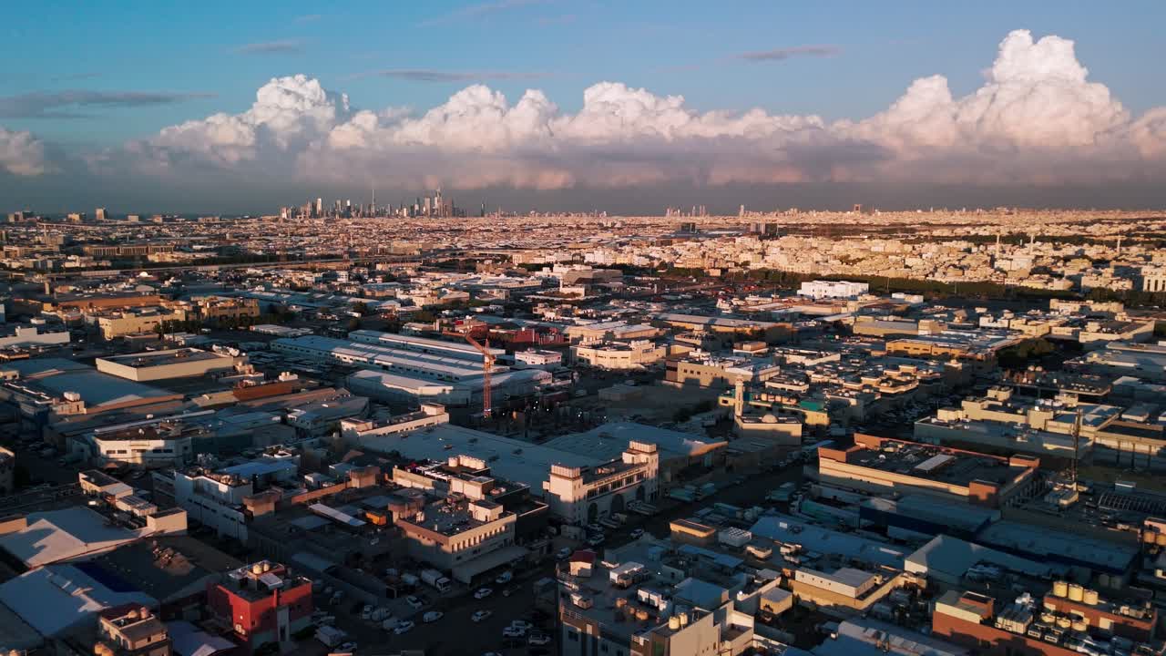 Ampia ripresa aerea con drone durante l'ora d'oro: skyline distante sotto una linea di cumulonembi, magazzini illuminati dal sole e griglia residenziale, cielo blu e lunghe ombre: pulito b-roll introduttivo per viaggi e uso aziendale