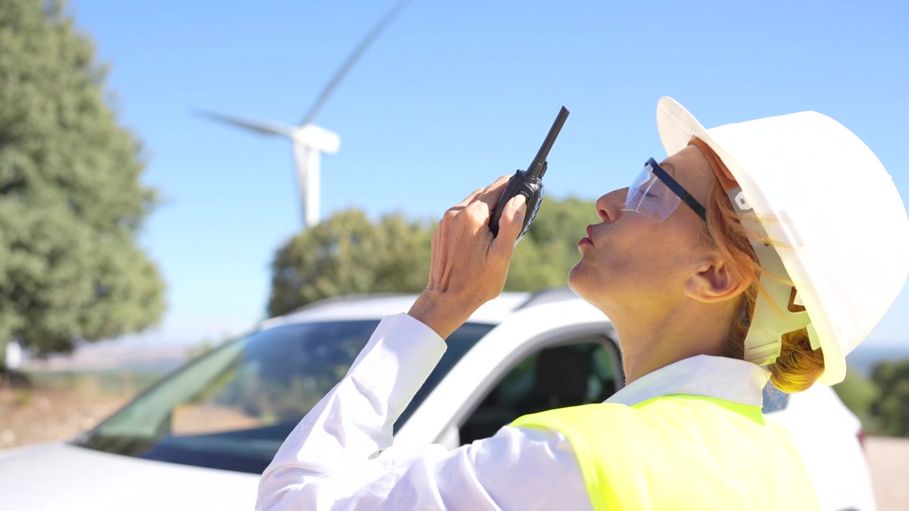 Woman in a hard hat using a walkie talkie at a renewable energy site with a wind turbine