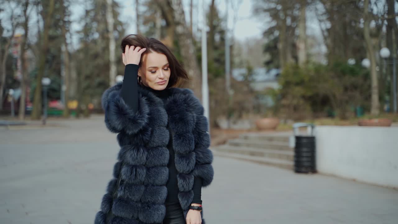 Street portrait of a fashionable woman. Young beautiful model wearing stylish black fur coat walking in the park. Model posing on camera. Street fashion concept.