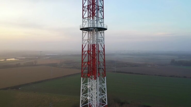 Un'alta torre di trasmissione metallica domina il primo piano, mentre campi rettangolari verdi e marroni si estendono verso un orizzonte nebbioso, con una luce calda e delicata e un leggero movimento della telecamera aerea.
