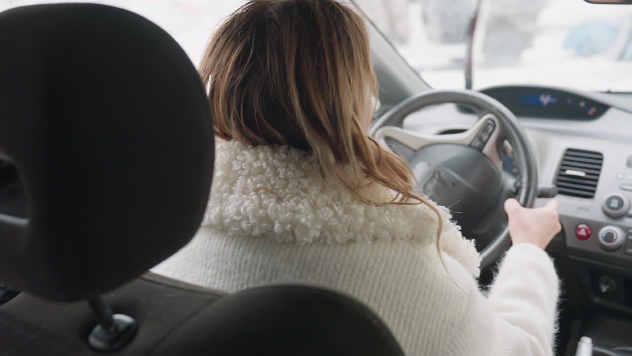 rear view of woman in white sweater holding steering wheel while reversing car, windshield covered with moisture, interior dashboard visible, soft natural light entering vehicle from outside garage